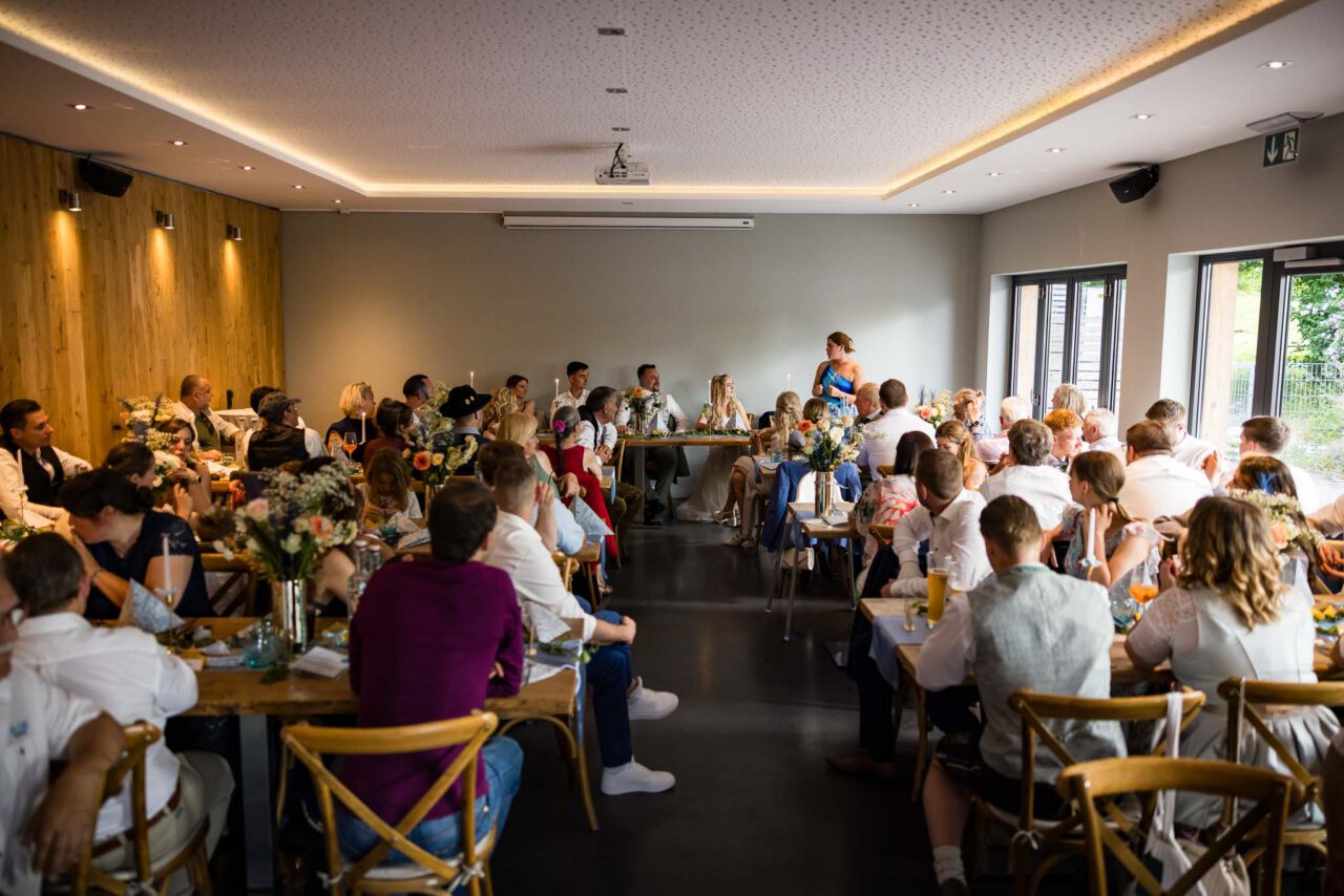 Das Brautpaar mit ihren Hochzeitsgästen in der Eventlocation im Saal während eine Rede gehalten wird