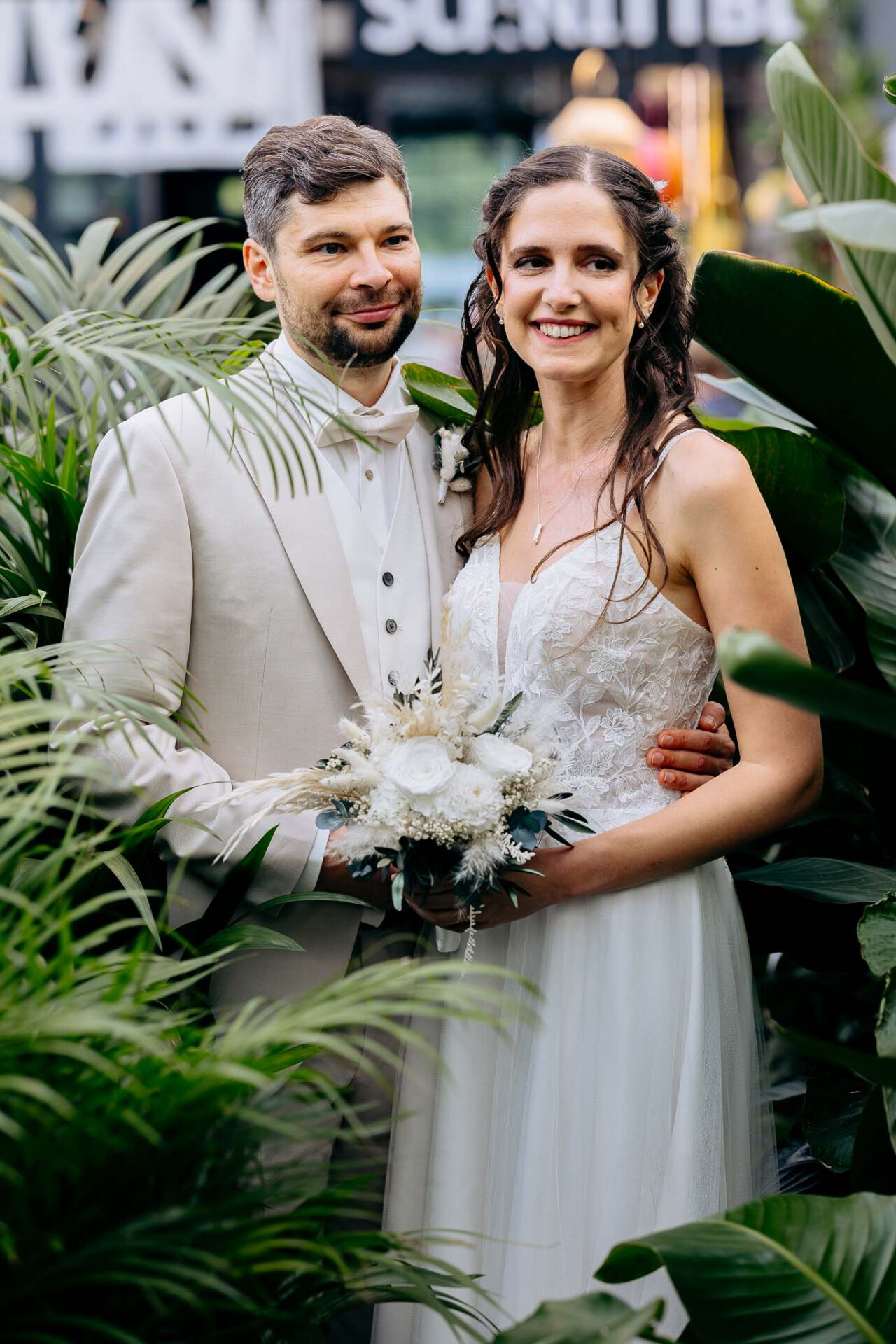 Zwischen den Pflanzen im Grünen steht ein Brautpaar im Gartencenter Kiefl lächelnd vor der Kamera