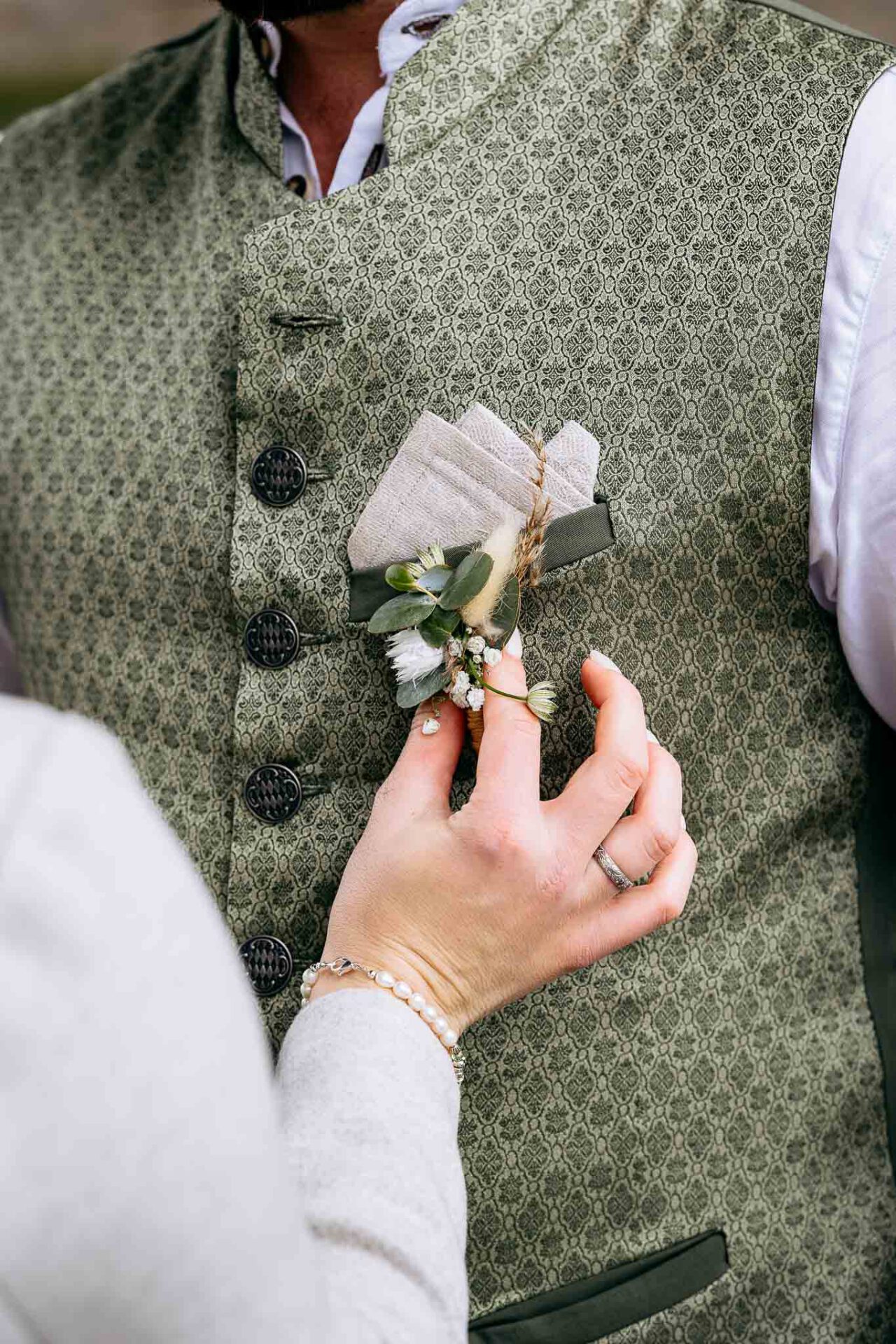 Die Braut richtet den Anstecker von ihrem Bräutigam an der Trachtenweste von Holledauer Herzens Gwand richtig hin