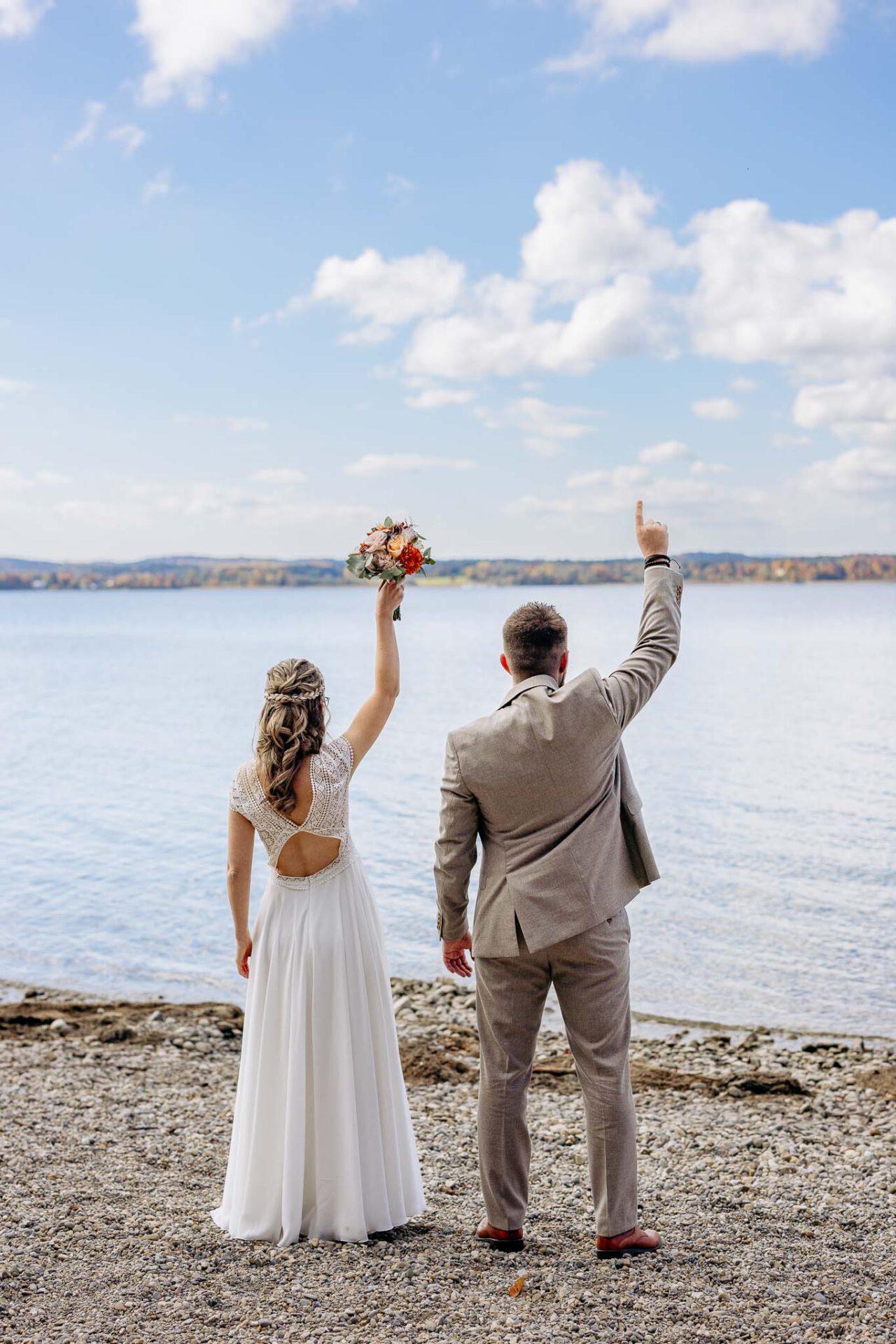 Die Braut reißt ihren Brautstrauß in die Luft und der Bräutigam zeigt mit dem Finger in den Himmel am Starnberger See