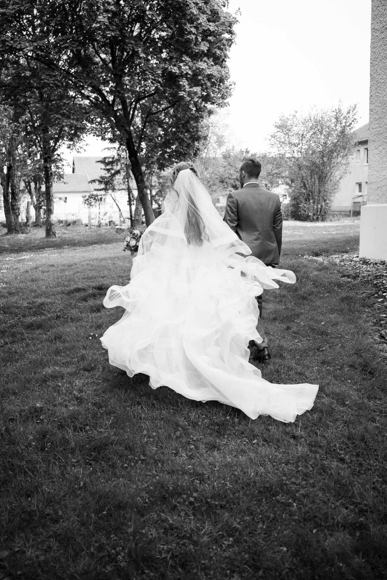 Schwarz Weiß Bild hinter der Kirche in Königsmoos mit dem Brautpaar im starken Wind das Brautkleid weh himmlisch hinter der Braut